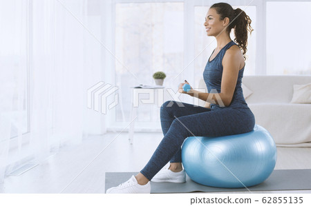 Attractive Hispanic lady with dumbbells sitting on fitness ball at home, copy space Attractive Hispanic lady with dumbbells sitting on fitness ball at home, copy space 62855135