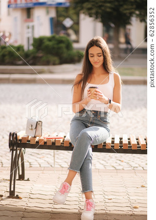 Attrective brunette sitting on the bench in centre of the city. Beautiful young woman ouside drink coffee Attrective brunette sitting on the bench in centre of the city. Beautiful young woman ouside drink coffee 62856352