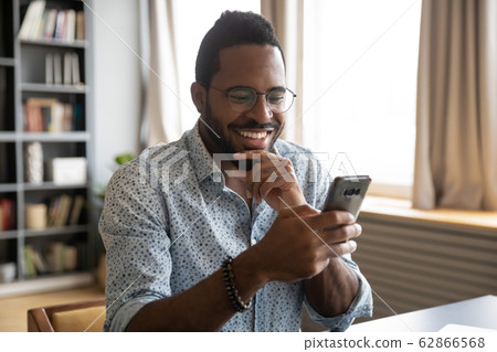Smiling millennial biracial guy reading pleasant good news sms. Smiling millennial biracial guy reading pleasant good news sms. 62866568