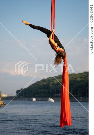 Beautiful and flexible female circus artist dancing with aerial silk with sky and river coastline on Beautiful and flexible female circus artist dancing with aerial silk with sky and river coastline on 62868441