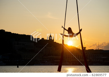 Silhouette of a flexible woman acrobat on aerial silk during a sunset on Kiev city background Silhouette of a flexible woman acrobat on aerial silk during a sunset on Kiev city background 62869577