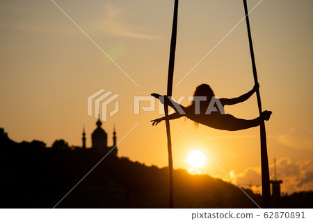 Silhouette of a flexible woman acrobat on aerial silk during a sunset on Kiev city background 62870891