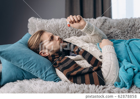Mature man lying on couch and looking at thermometer Mature man lying on couch and looking at thermometer 62878858