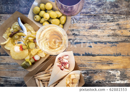 Tapas, pinchos, and wine. Cheese, jamon and olives, spicy gildas and potato chips, shot from the top on a wooden table in an outdoors cafe with a place for text Tapas, pinchos, and wine. Cheese, jamon and olives, spicy gildas and potato chips, shot from the top on a wooden table in an outdoors cafe with a place for text 62881553