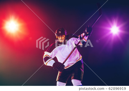 Male hockey player with the stick on ice court and dark neon colored background Male hockey player with the stick on ice court and dark neon colored background 62896919