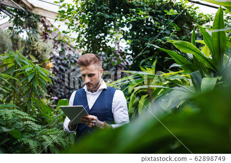Young man with tablet standing in botanical garden, working. 62898749