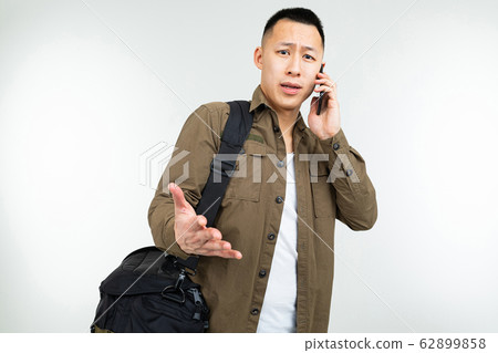 Asian man in urban clothes with a bag communicates using a smartphone about a business on a white Asian man in urban clothes with a bag communicates using a smartphone about a business on a white 62899858
