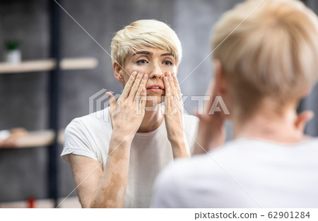 Tired Middle-Aged Woman Touching Face Looking In Mirror In Bathroom 62901284