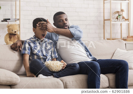 Father and son watching scary movie together, dad covering kid's eyes Father and son watching scary movie together, dad covering kid's eyes 62901886