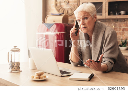 Concerned elderly woman talking on cellphone, working on laptop in kitchen Concerned elderly woman talking on cellphone, working on laptop in kitchen 62901893