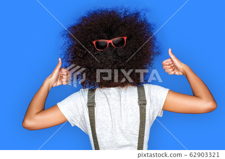 Having fun. Mulatto girl standing isolated on blue back view wearing sunglasses on hair playful thumbs up Having fun. Mulatto girl standing isolated on blue back view wearing sunglasses on hair playful thumbs up 62903321
