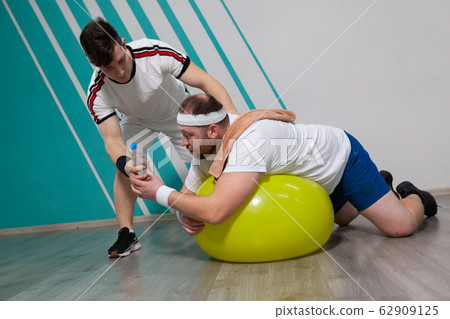 Overweight man is lying on the fitness ball in the gym while his personal trainer offers him a bottle of water. Fat man is dying of thirst and looking exhausted after a hard training in fitness group Overweight man is lying on the fitness ball in the gym while his personal trainer offers him a bottle of water. Fat man is dying of thirst and looking exhausted after a hard training in fitness group 62909125