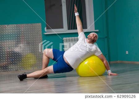 Smiling fat man is doing exercises using fitness ball. Overweight man is happy with the result of his training in group fitness classes Smiling fat man is doing exercises using fitness ball. Overweight man is happy with the result of his training in group fitness classes 62909126