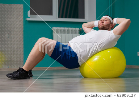 Overweight man is lying on a fitness ball during group fitness classes. Fat man looks disappointed because of bad result of weight loss trainings Overweight man is lying on a fitness ball during group fitness classes. Fat man looks disappointed because of bad result of weight loss trainings 62909127