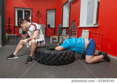 Personal trainer is dissatisfied with his overweight sportsman who is lying exhausted with his head into a tire after hard training in the gym Personal trainer is dissatisfied with his overweight sportsman who is lying exhausted with his head into a tire after hard training in the gym 62909140