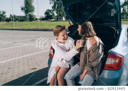 Two girls in the parking lot at the open trunk posing for the camera. Two girls in the parking lot at the open trunk posing for the camera. 62911443