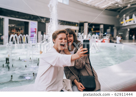 Two girls take a selfie in the mall, a fountain in the background Two girls take a selfie in the mall, a fountain in the background 62911444