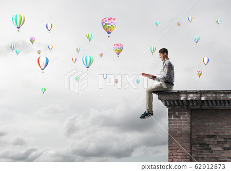 Man on roof edge reading book and aerostats flying in sky Man on roof edge reading book and aerostats flying in sky 62912873