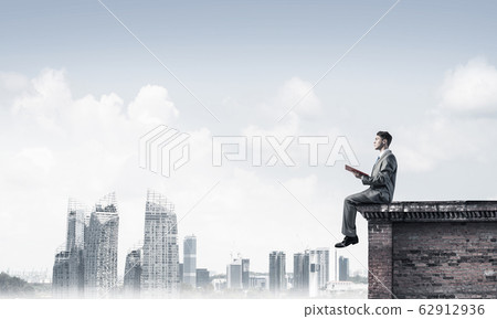 Man on roof edge reading book and cityscape at background Man on roof edge reading book and cityscape at background 62912936