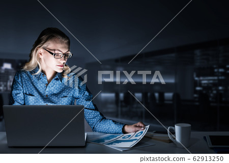 Attractive blonde working on laptop in dark office. Mixed media 62913250