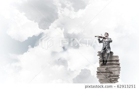 Handsome violinist in blue day sky on pile of books play his mel 62913251