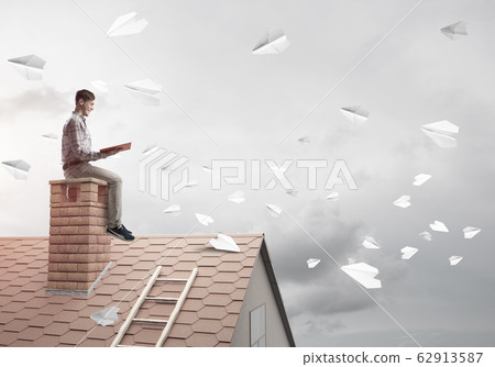 Man on brick roof reading book and paper planes flying in air Man on brick roof reading book and paper planes flying in air 62913587