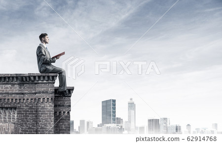Man on roof edge reading book and cityscape at background Man on roof edge reading book and cityscape at background 62914768