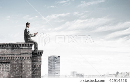 Man on roof edge send message with smartphone and cityscape at b Man on roof edge send message with smartphone and cityscape at b 62914838