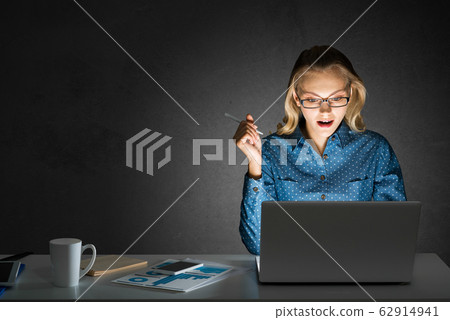 Attractive blonde working on laptop in dark office. Mixed media 62914941