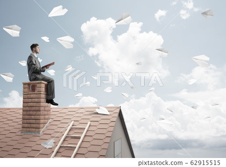 Man on brick roof reading book and paper planes flying in air Man on brick roof reading book and paper planes flying in air 62915515
