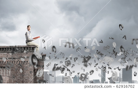Man on roof edge reading book and symbols flying around Man on roof edge reading book and symbols flying around 62916702