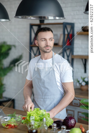 Dark-haired man in a kitchen apron cooking at home. Dark-haired man in a kitchen apron cooking at home. 62922157
