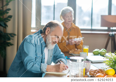 Bearded grey-haired man sitting at the table and making notes Bearded grey-haired man sitting at the table and making notes 62924053