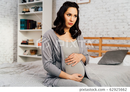 Asian mixed Caucasian anxious woman in pregnancy overthinking sits on bed at home Asian mixed Caucasian anxious woman in pregnancy overthinking sits on bed at home 62925662