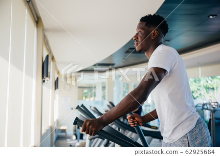 Side view of fit African American man in sportswear doing steps on elliptical machine during workout in modern gym 62925684