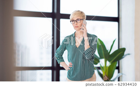 Strict Businesswoman Talking On Phone Standing Near Window In Office Strict Businesswoman Talking On Phone Standing Near Window In Office 62934410