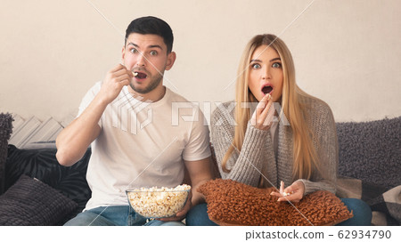 Surprised young couple with popcorn watching TV in living room 62934790