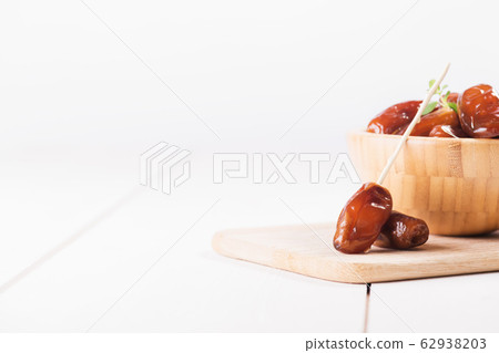 Close up of a date on a stick. Raw sweet dry dates with in a wooden bowl on a white background. Organic sweeties to healthy eating. Close up of a date on a stick. Raw sweet dry dates with in a wooden bowl on a white background. Organic sweeties to healthy eating. 62938203