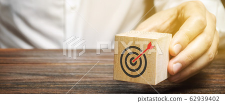 A businessman holds in his hands a wooden block with a target symbol. The concept of achieving business goals. Execution of a business plan. Purposefulness. Goal. Planning. Strategy A businessman holds in his hands a wooden block with a target symbol. The concept of achieving business goals. Execution of a business plan. Purposefulness. Goal. Planning. Strategy 62939402