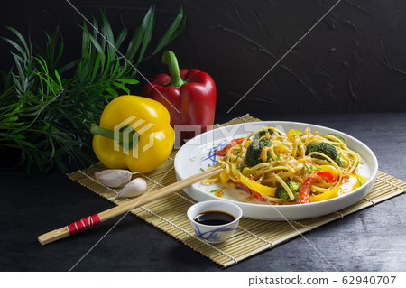 Udon noodles with vegetables on a black plate with soy sauce and ingredients on a bamboo mat, black background Udon noodles with vegetables on a black plate with soy sauce and ingredients on a bamboo mat, black background 62940707