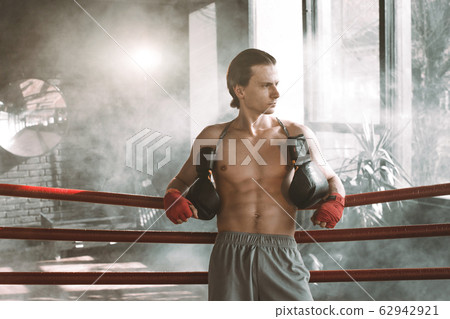 Male boxer having muscular body standing inside a boxing ring. Boxer resting his arms on the rope of boxing ring. Male boxer having muscular body standing inside a boxing ring. Boxer resting his arms on the rope of boxing ring. 62942921