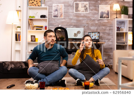 Couple sitting on the floor and watching TV in their living room Couple sitting on the floor and watching TV in their living room 62956840