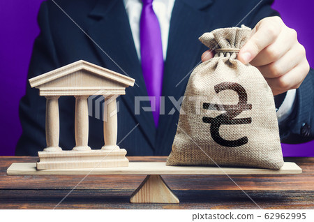 A man puts a ukrainian hryvnia money bag on scales opposite to government building or bank. State debt. Budget balance deficit. Taxes payment. Lobbying interests. Monetary policy. High corruption risk 62962995