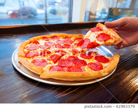 Just cooked Pizza pepperoni on a table in a cafe in front of a window with a panorama of the city. Appetizing hot pizza with slices of smoked sausage. A man tears a slice of pizza chopped into pieces Just cooked Pizza pepperoni on a table in a cafe in front of a window with a panorama of the city. Appetizing hot pizza with slices of smoked sausage. A man tears a slice of pizza chopped into pieces 62963379