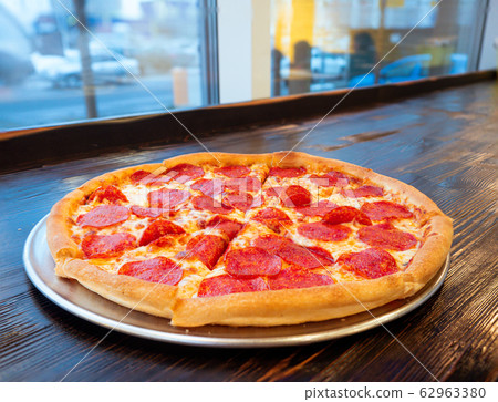 Just cooked Pizza pepperoni on a table in a cafe in front of a window with a panorama of the city. Appetizing hot pizza with slices of smoked sausage Just cooked Pizza pepperoni on a table in a cafe in front of a window with a panorama of the city. Appetizing hot pizza with slices of smoked sausage 62963380