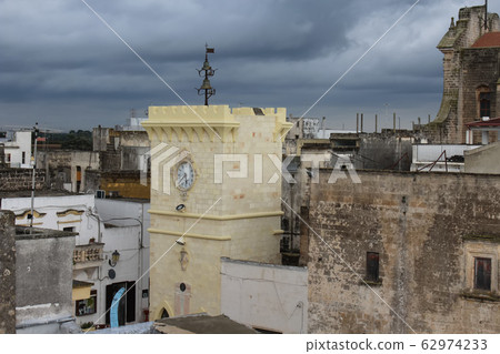 Avetrana, Italy - October, 10, 2019, Clock tower in tiny Italian paese Avetrana, province of Taranto, Puglia, southern Italy. Autumn cloudy day with blue clouds. View from the roof. Medieval heritage. 62974233