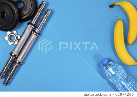 Fitness or bodybuilding. Sports equipment on a blue background, banana, water bottle, barbell, dumbbell, top view with plenty of copy space Fitness or bodybuilding. Sports equipment on a blue background, banana, water bottle, barbell, dumbbell, top view with plenty of copy space 62974296