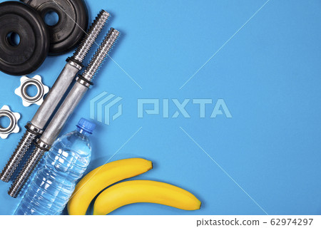 Fitness or bodybuilding. Sports equipment on a blue background, banana, water bottle, barbell, dumbbell, top view with plenty of copy space Fitness or bodybuilding. Sports equipment on a blue background, banana, water bottle, barbell, dumbbell, top view with plenty of copy space 62974297