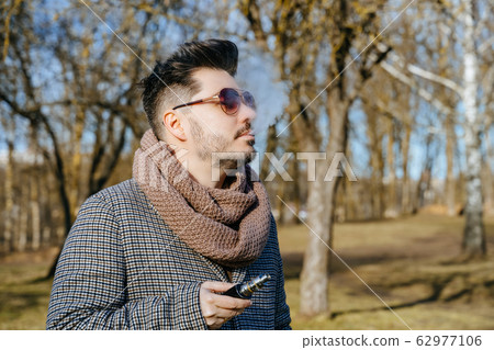Confident young bearded male in sunglasses vape electronic cigarette. A young handsome bearded hipster man vaping e-cigarette outdoors in the park. Close up. 62977106