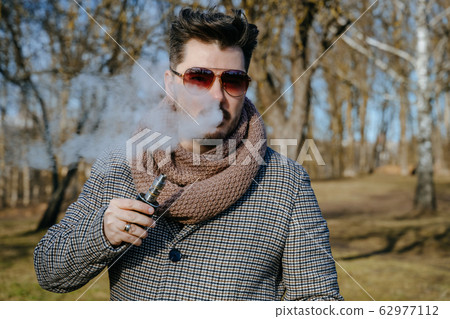 Confident young bearded male in sunglasses vape electronic cigarette. A young handsome bearded hipster man vaping e-cigarette outdoors in the park. Close up. 62977112